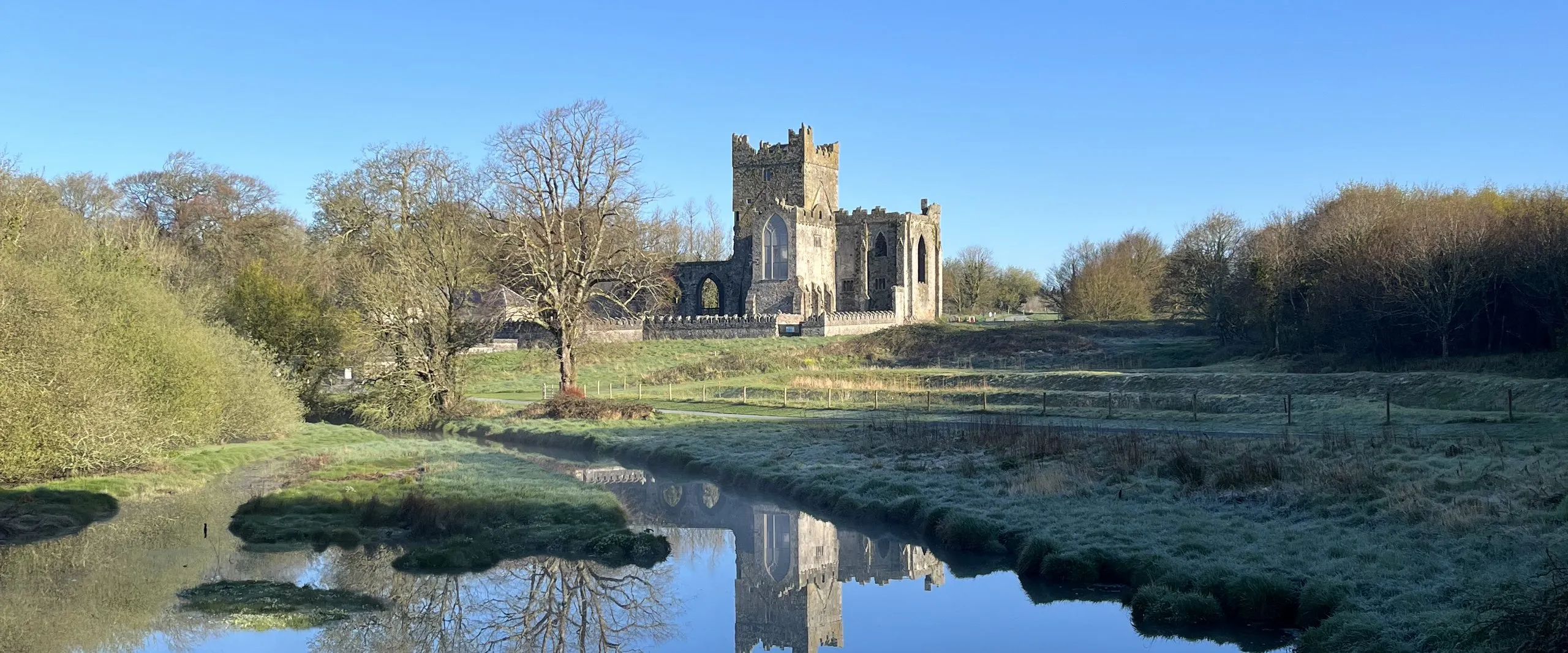 Tintern Abbey mirrored in the still river on a clear morning, the medieval tower and bare trees reflected in the blue water, frost on the far bank.