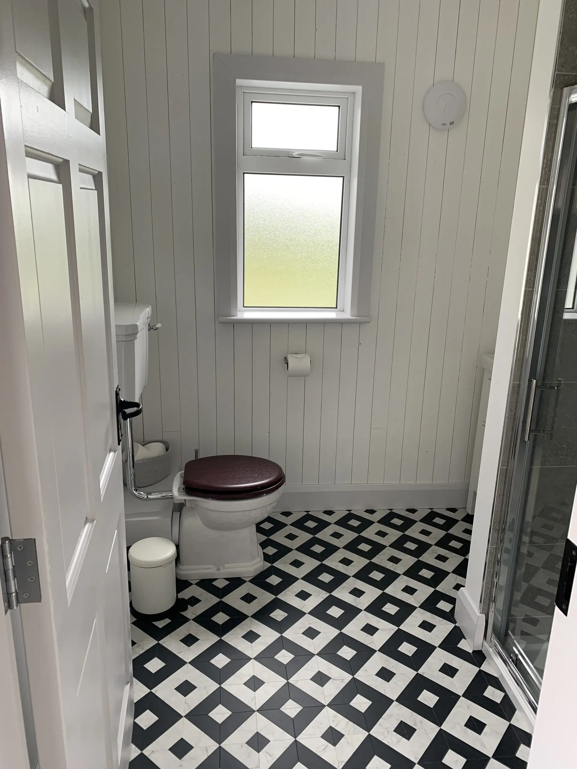A bathroom with patterned black-and-white floor tiles, white panelled walls, toilet and a glazed shower.