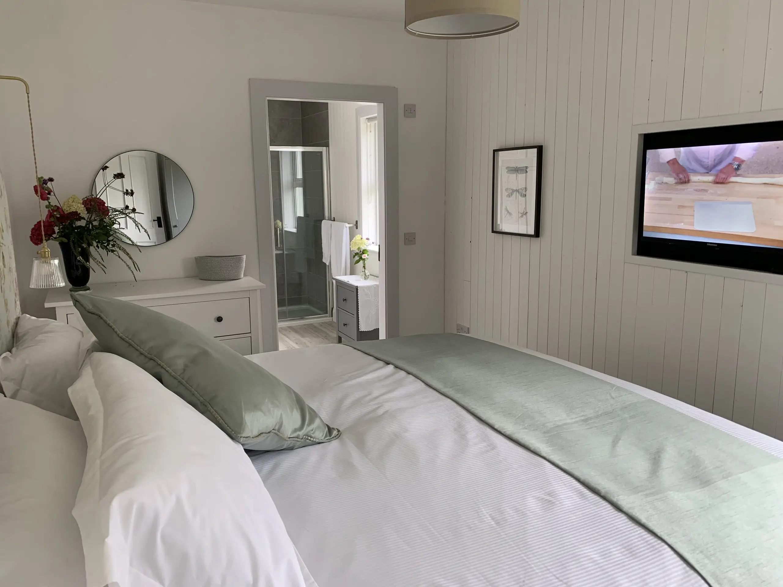 The Paddock Bedroom from the doorway, bed made up in white and sage green, with a round mirror and framed art.