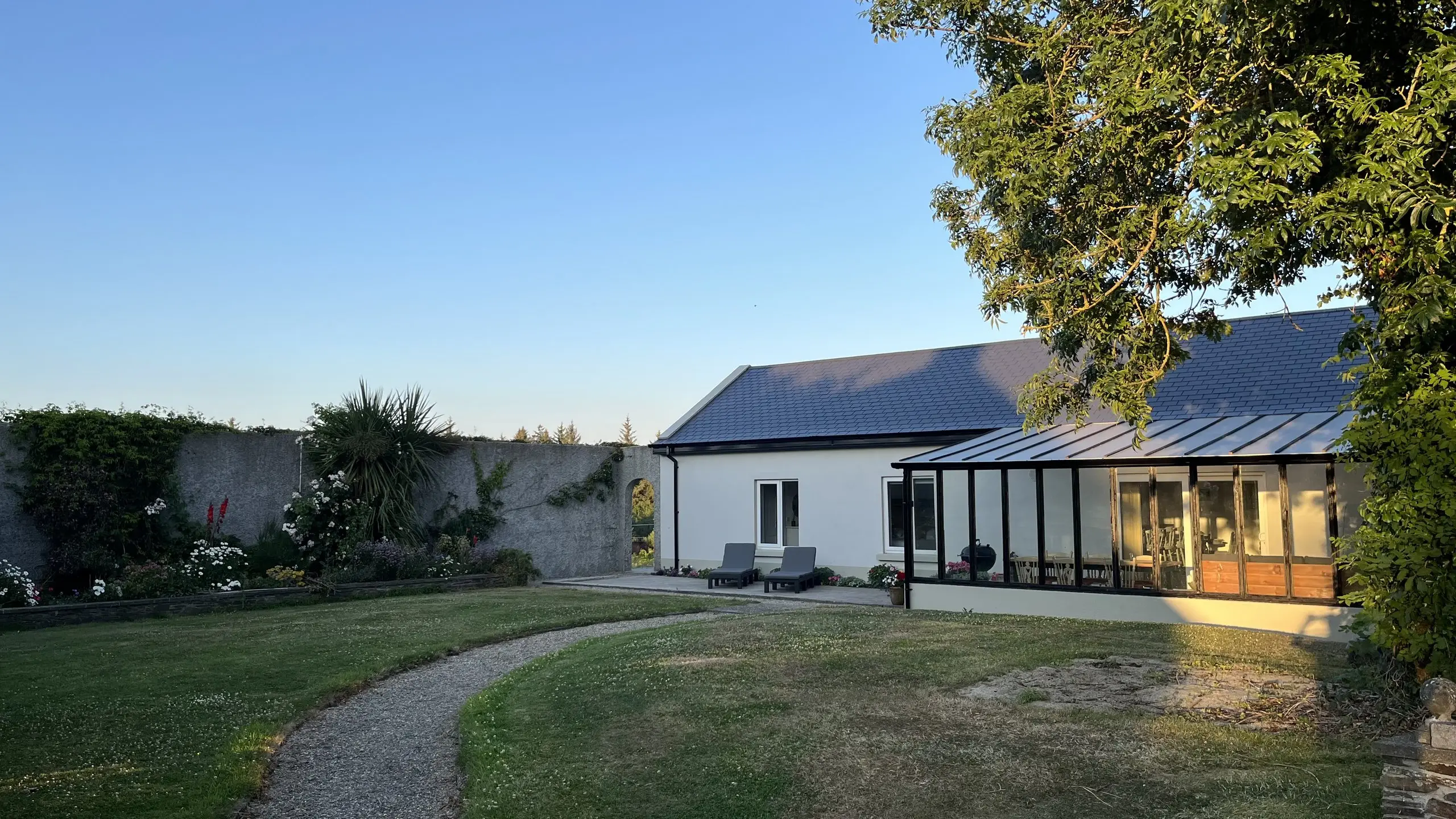 The cottage on a summer evening, the whole white building with its glazed veranda lit from inside and a gravel path curving across the lawn.