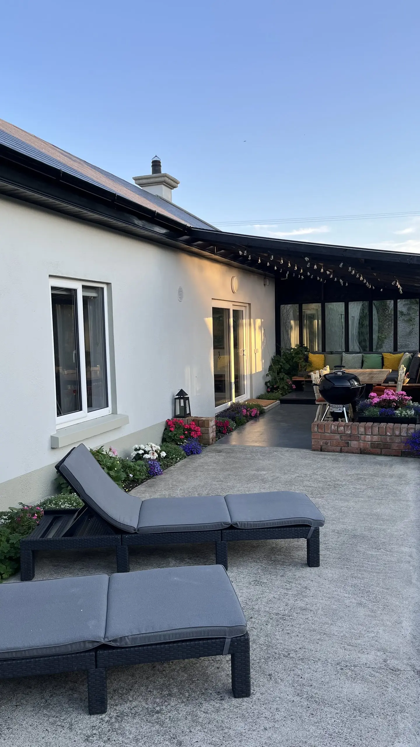 The terrace at dusk with sun loungers, a lit lantern, flower pots and a barbecue, the porch glowing behind.