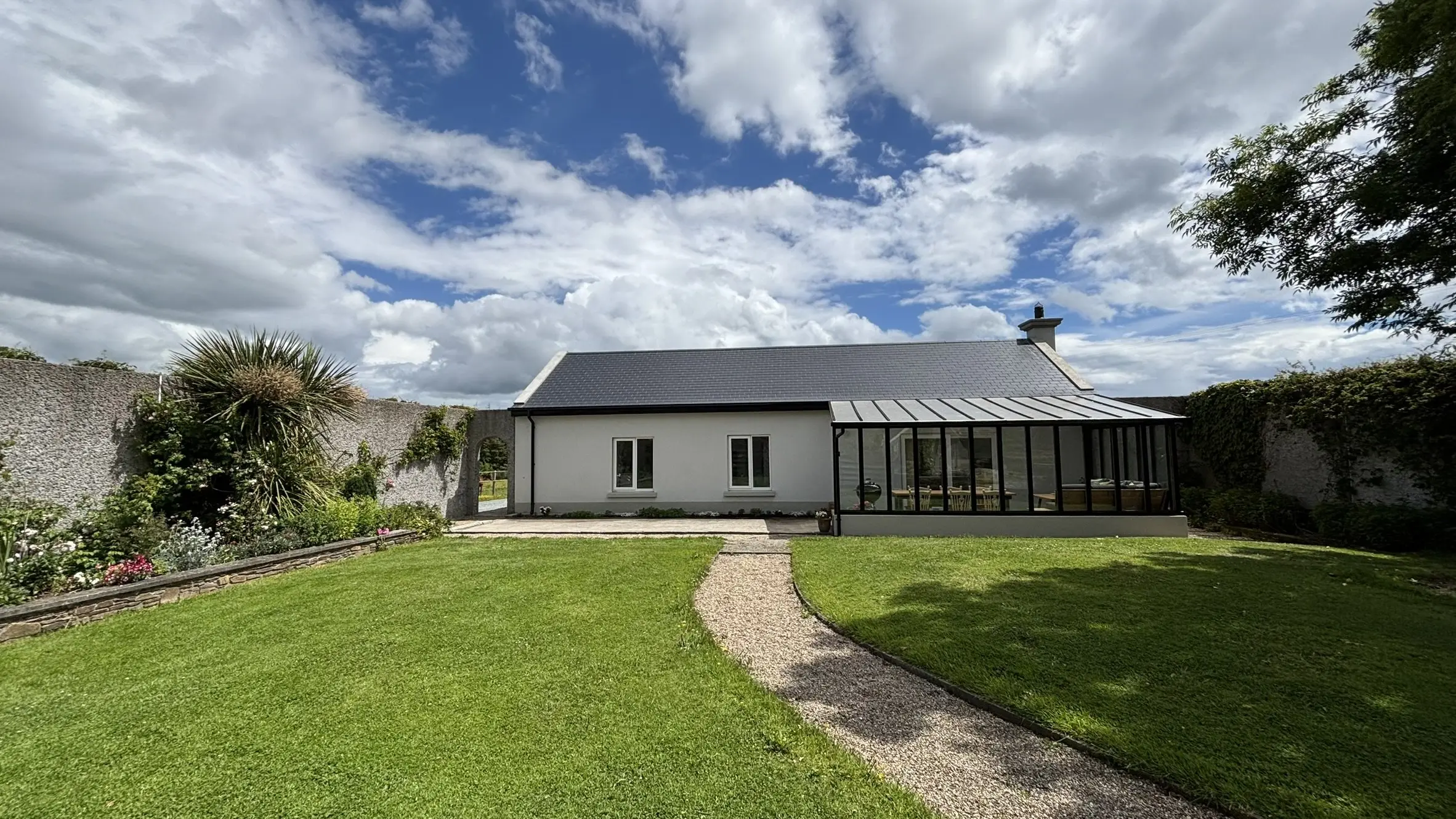 The cottage in daylight under a big cloudy sky, the long white building with its glazed veranda and lawn sweeping in front.