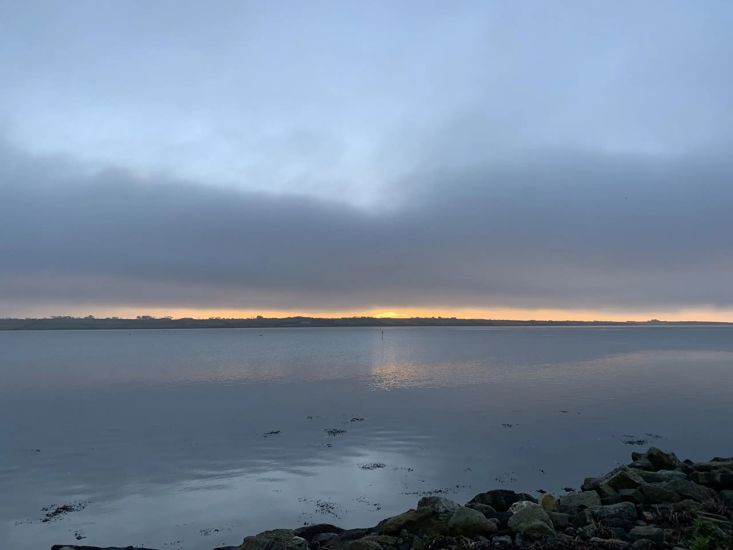 Dawn over Bannow Bay, a band of golden light along the horizon above the calm grey water, rocks in the foreground.