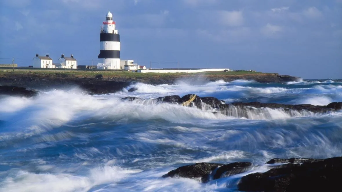 Hook Head lighthouse, black and white banded, standing over waves breaking on a rocky shore.