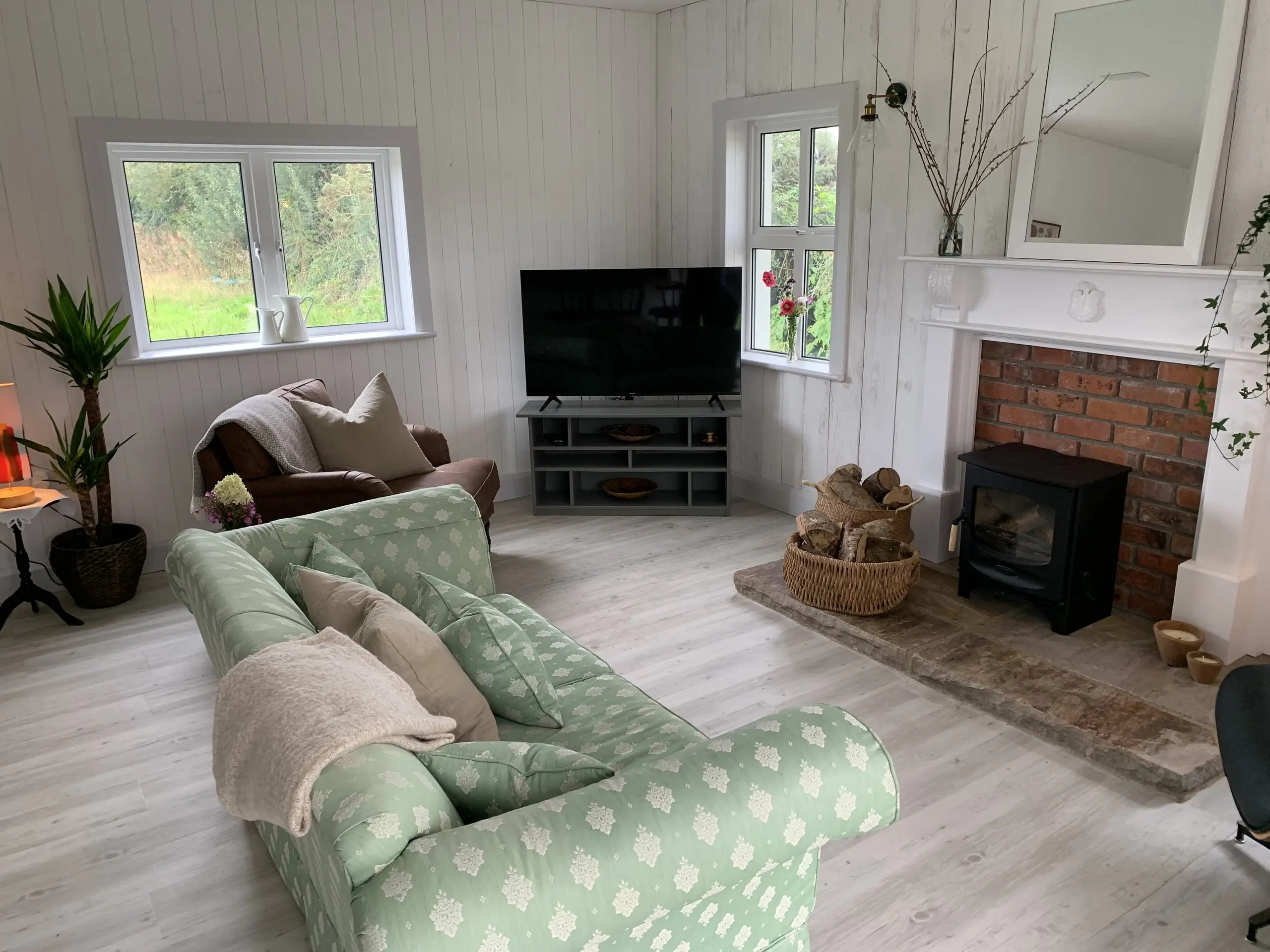 The living area with the wood stove, a green patterned sofa and armchair drawn up to it, and a basket of logs beside the brick fireplace.