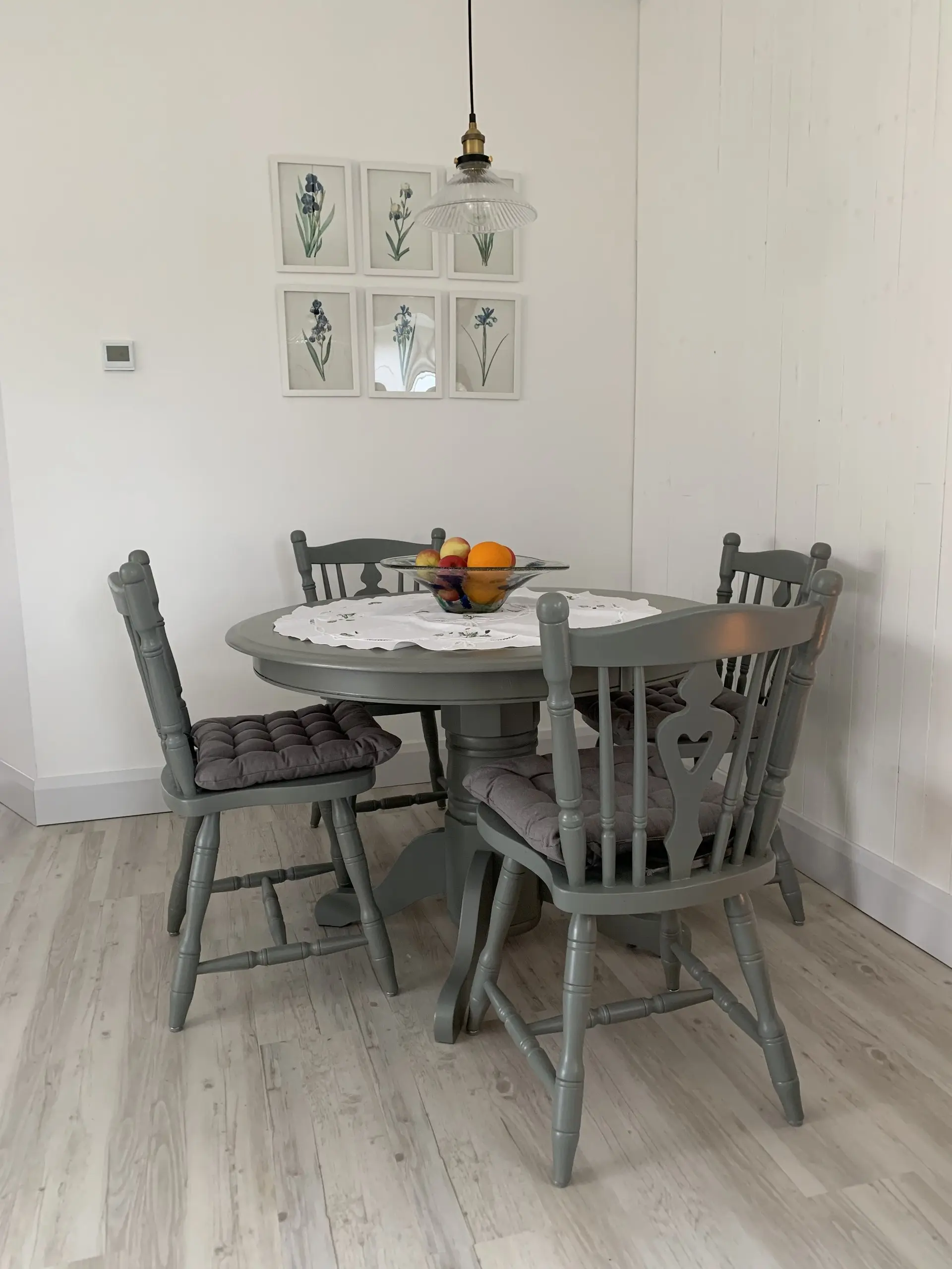 The grey round dining table and four chairs, with a bowl of fruit on top, against white panelled walls.