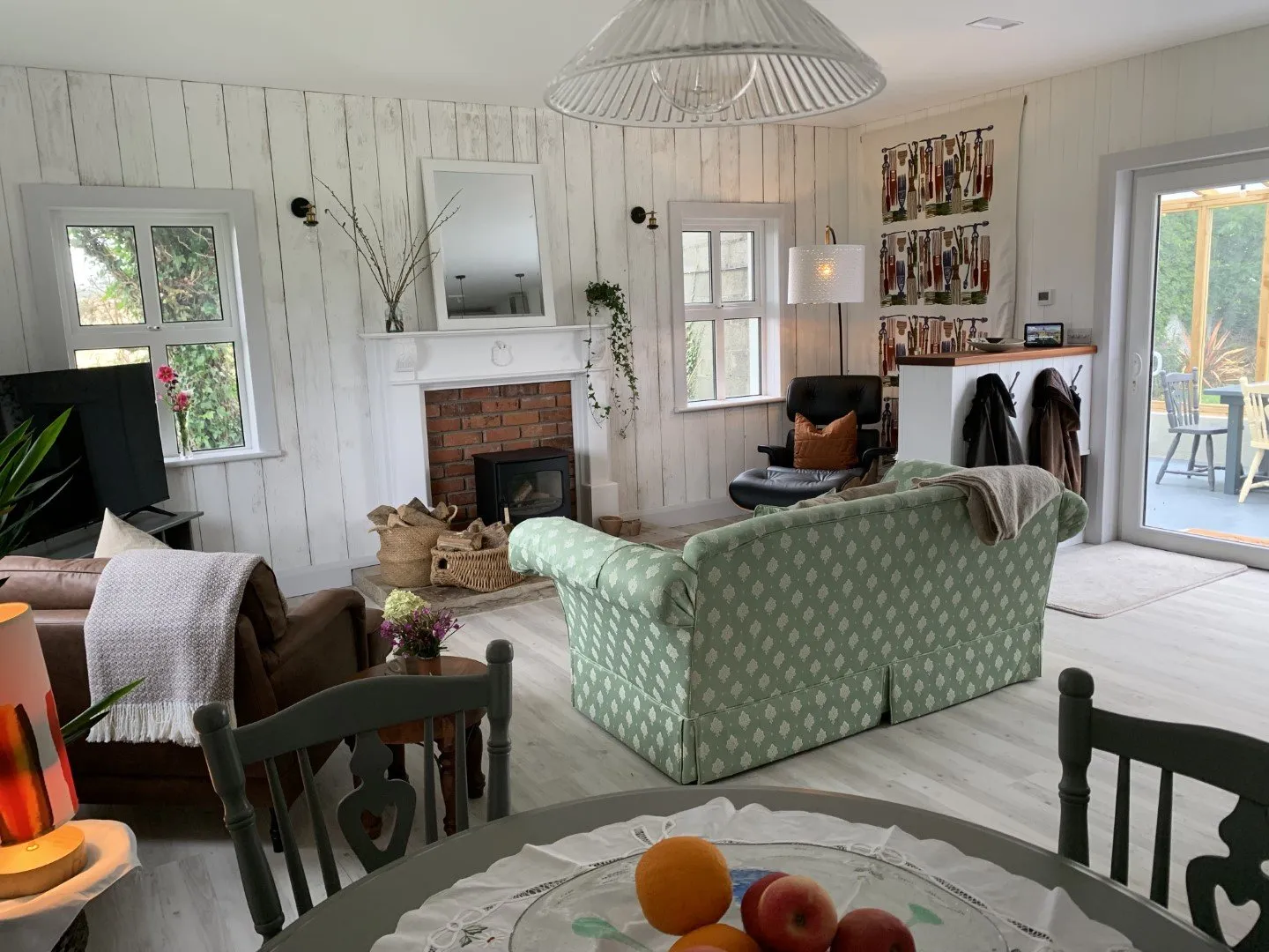 The open-plan living area: a green patterned sofa and armchairs around a fireplace, with a dining table and doors onto the garden.