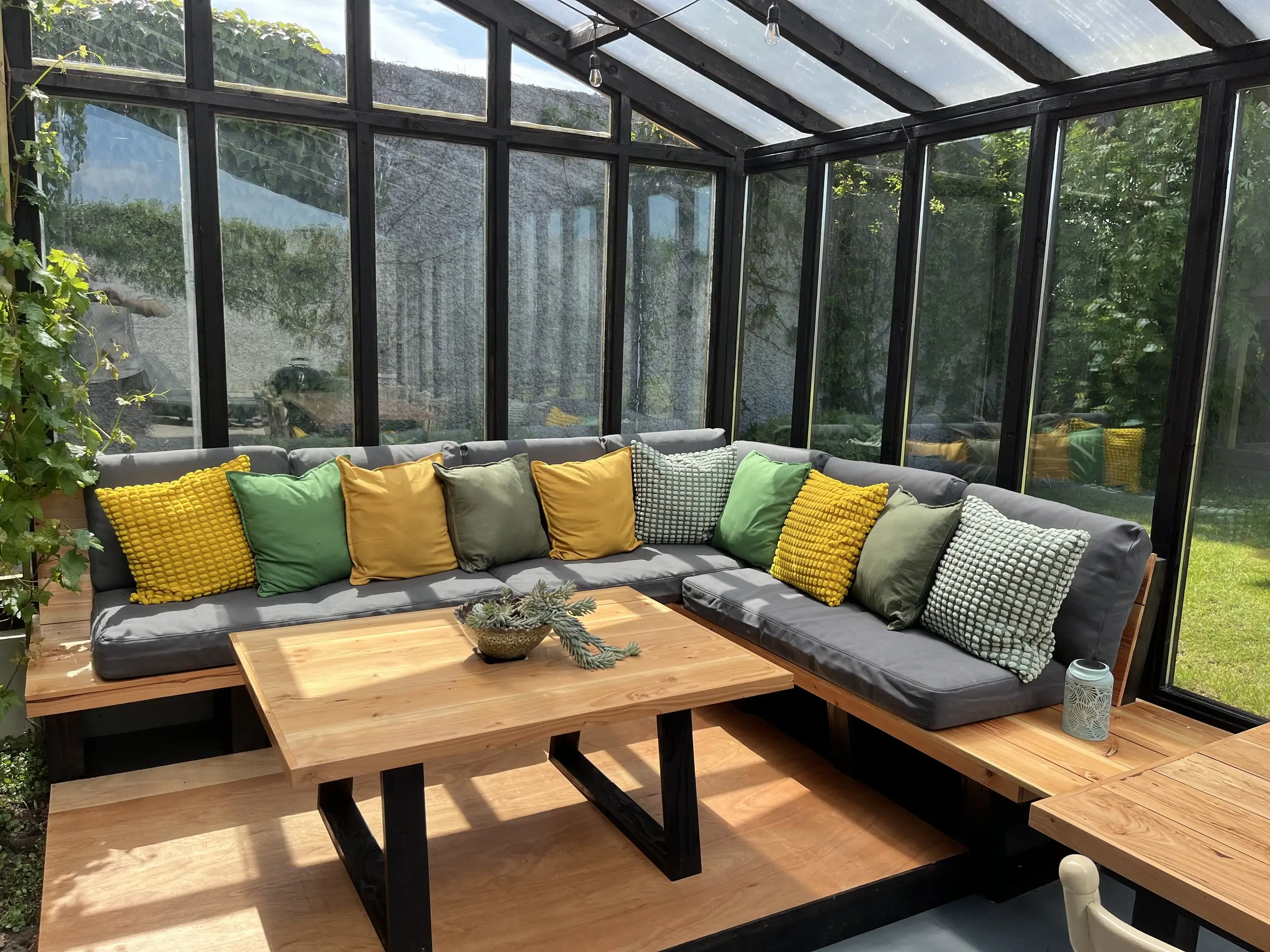 The glazed porch set for dining: a long oak table with yellow and green cushioned seating, a grapevine along the glass and a kettle barbecue.