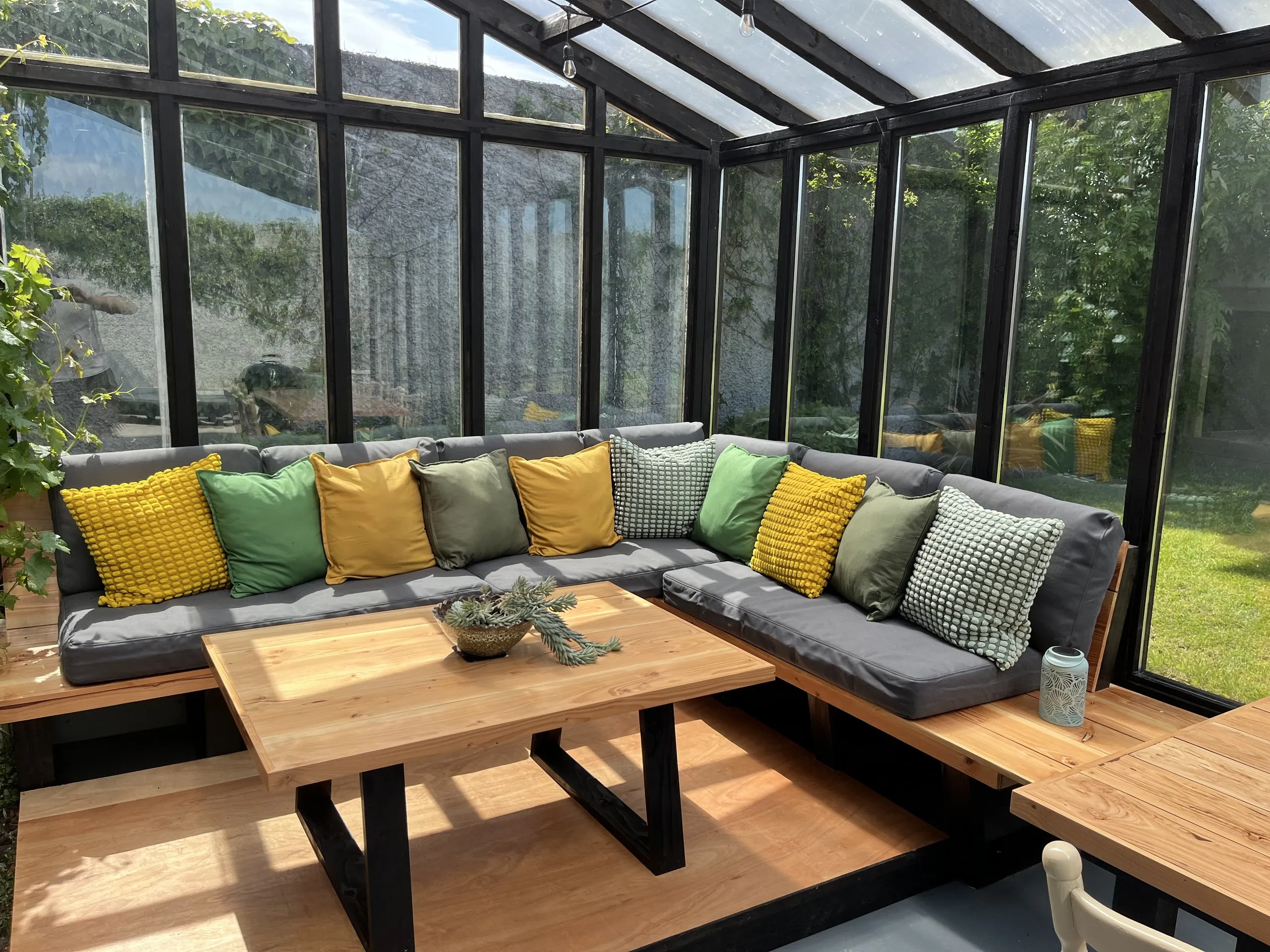The glazed porch: a large corner sofa banked with yellow and green cushions around an oak coffee table, with sunlight through the glass roof and the garden on every side.