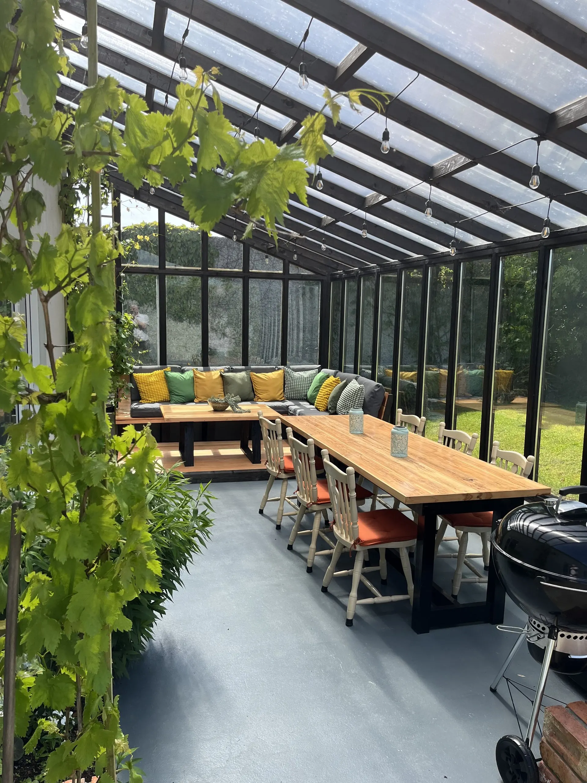 The length of the porch, with a grapevine growing along the glass and a long dining table beside the built-in bench seating.