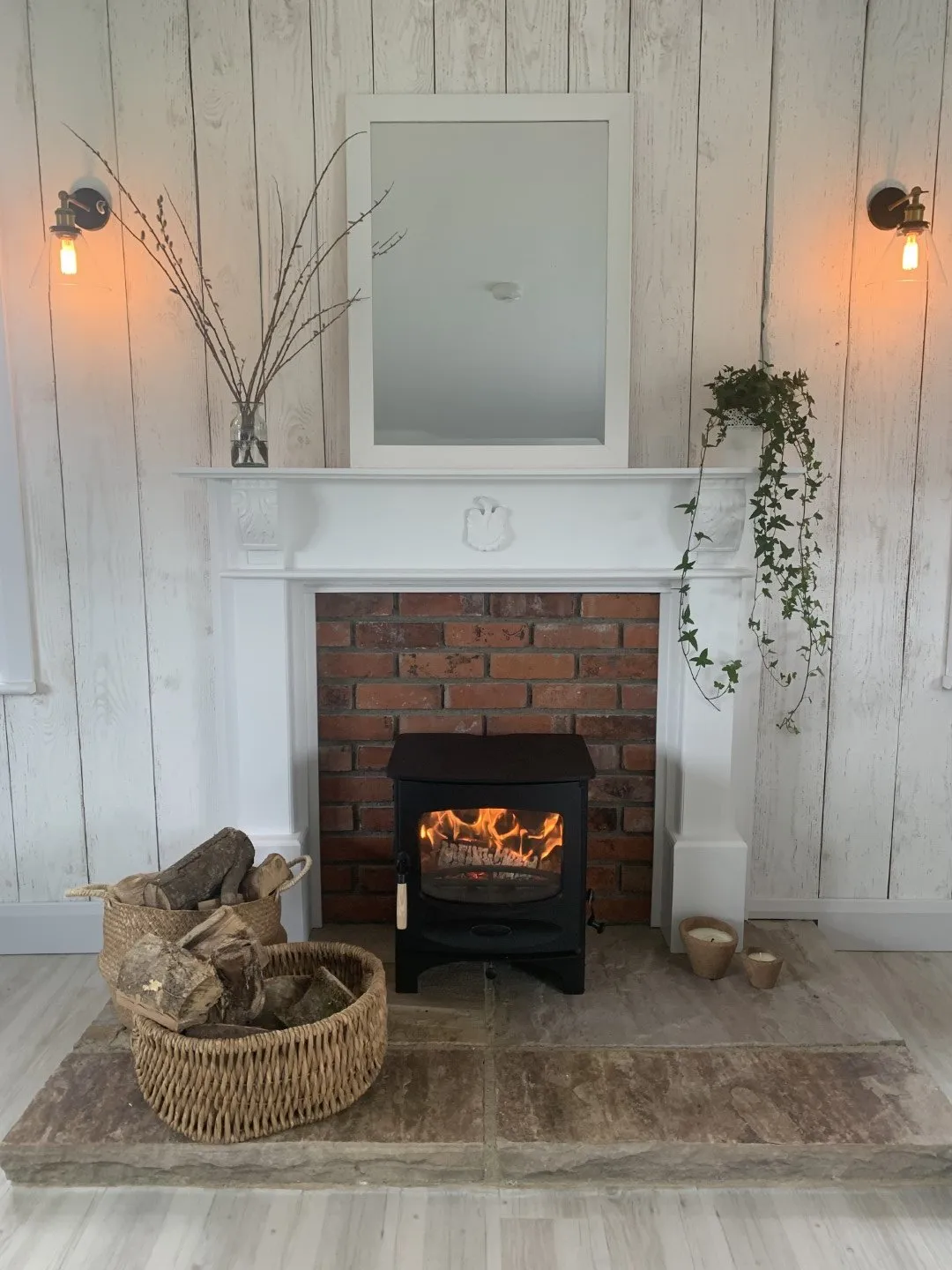 A lit wood-burning stove in a brick fireplace with a white mantel, a basket of logs beside it and trailing ivy above.